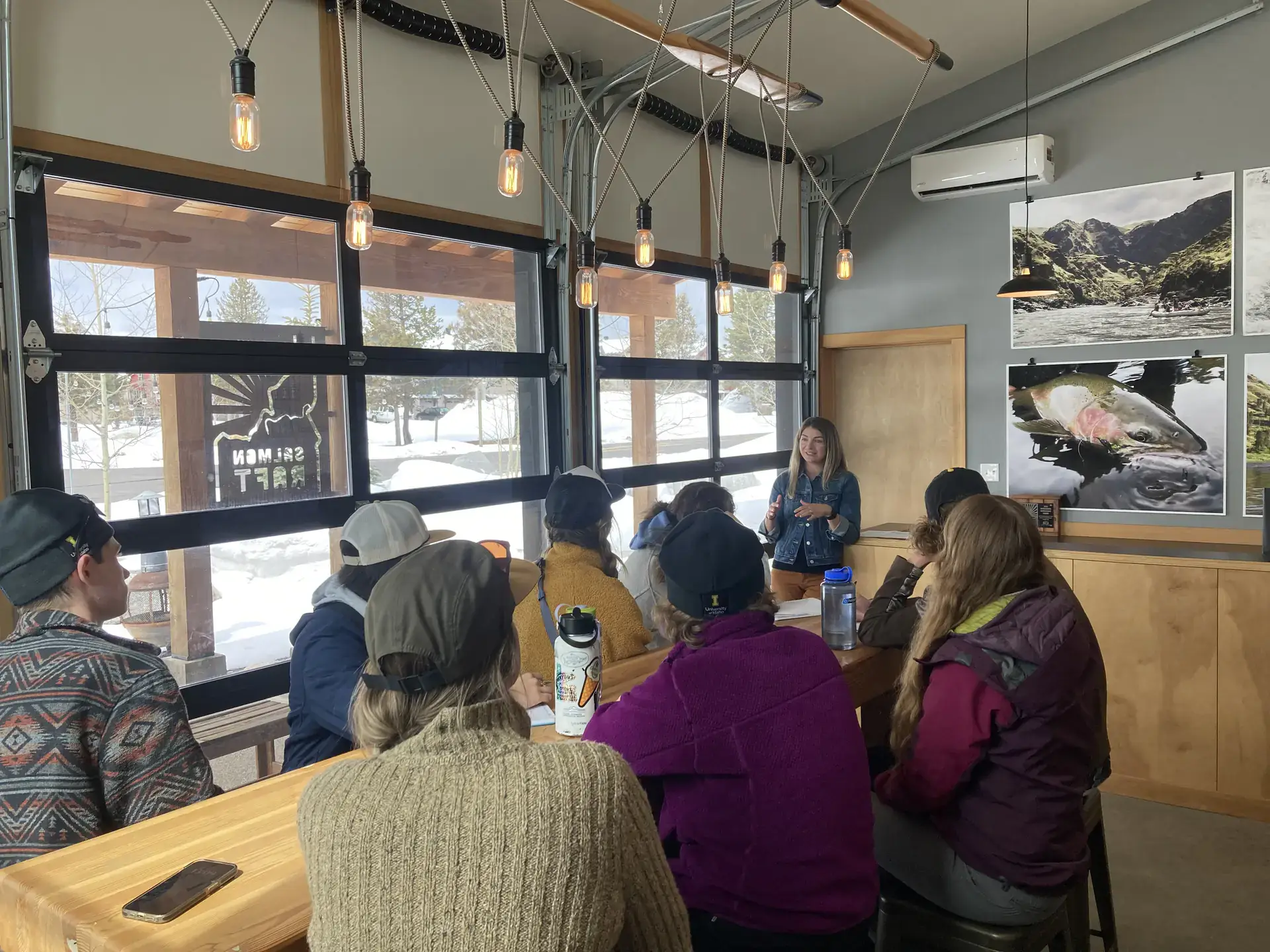 A group of students listen to a woman speaking inside a room filled with pictures of salmon and nature landscapes.