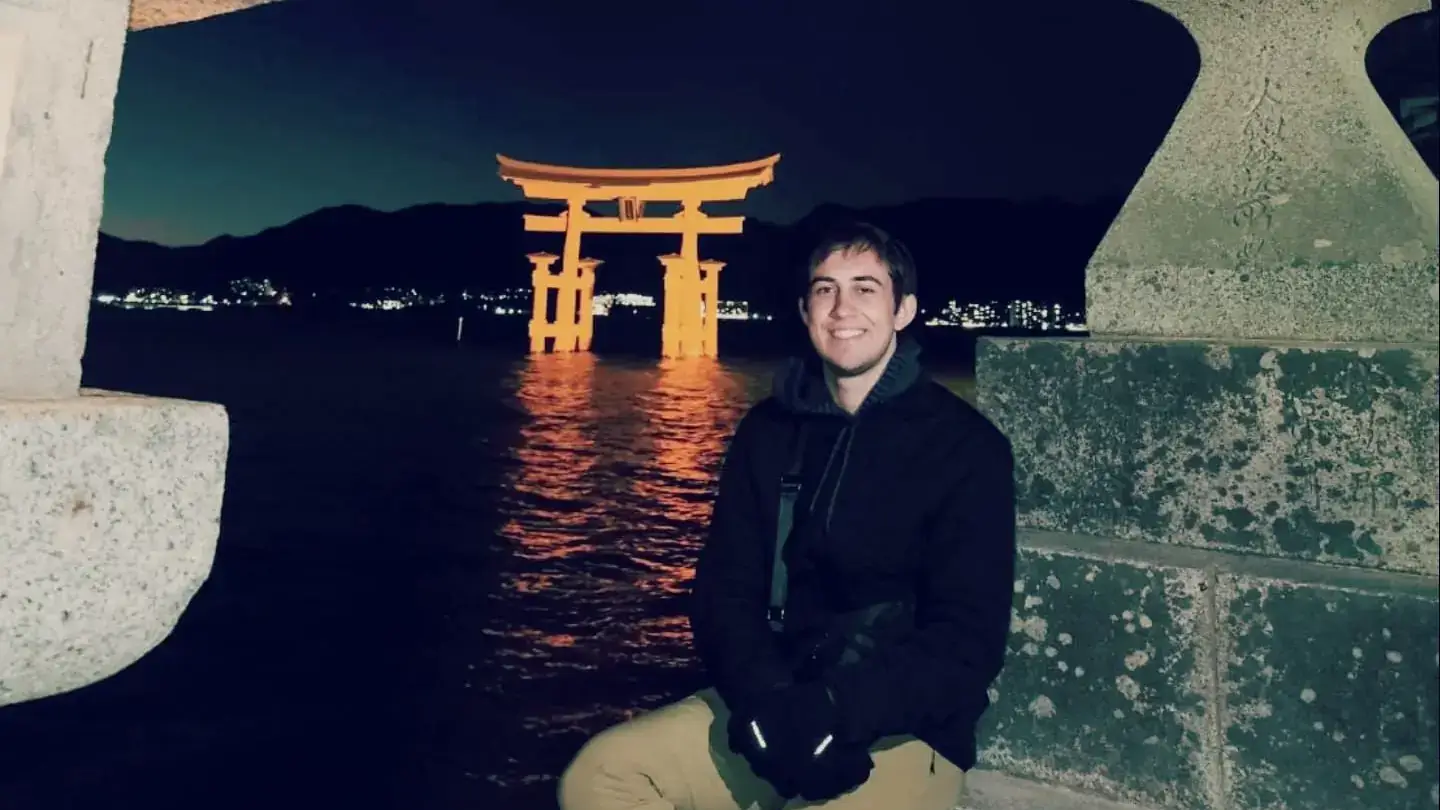 Shane Neirinckx smiles and sits on a concrete pillar at night with a Japanese torii behind him.