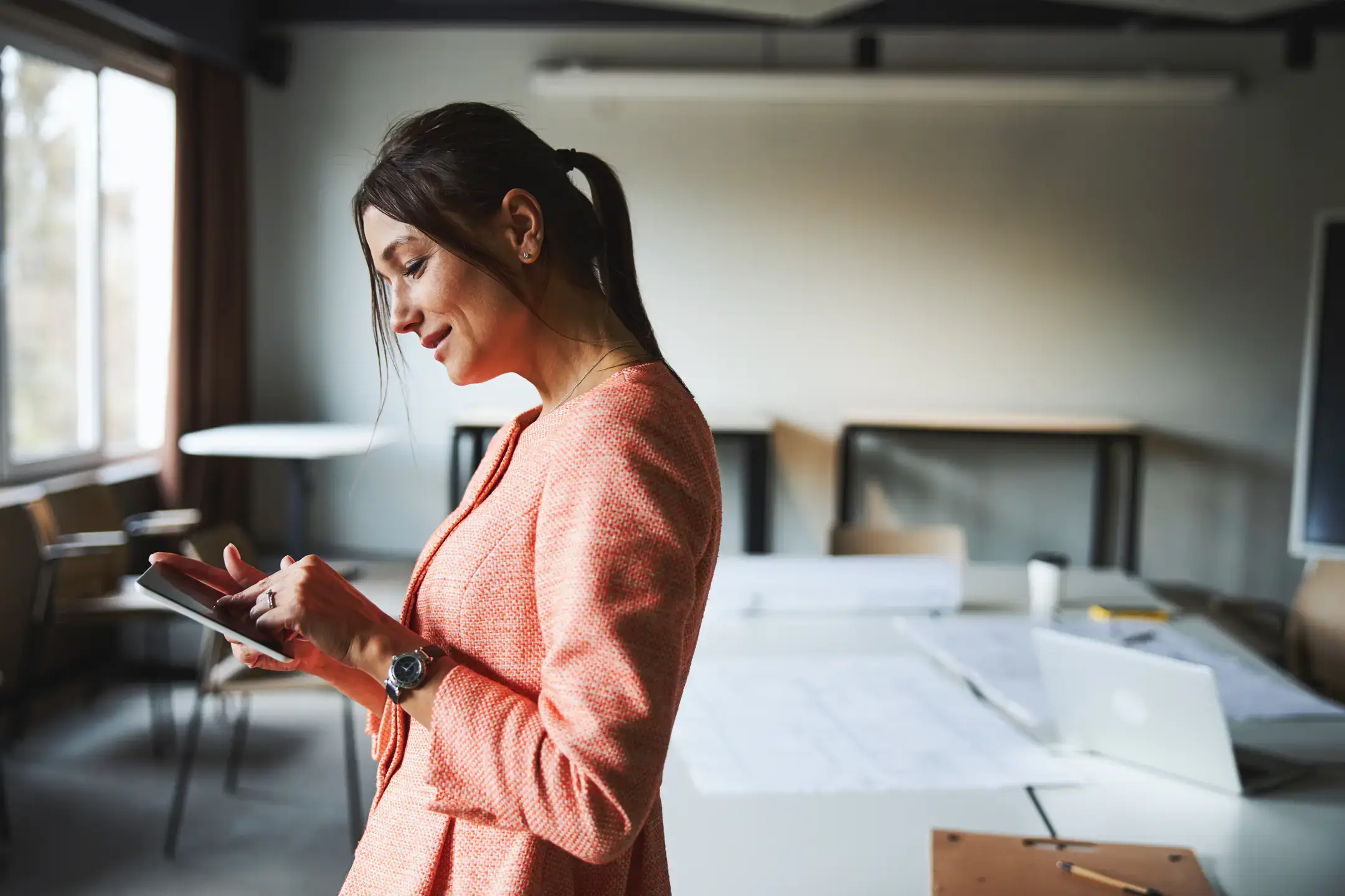 Young attractive Caucasian business woman in pink jacket holding tablet in hands while standing in the modern bright meeting room