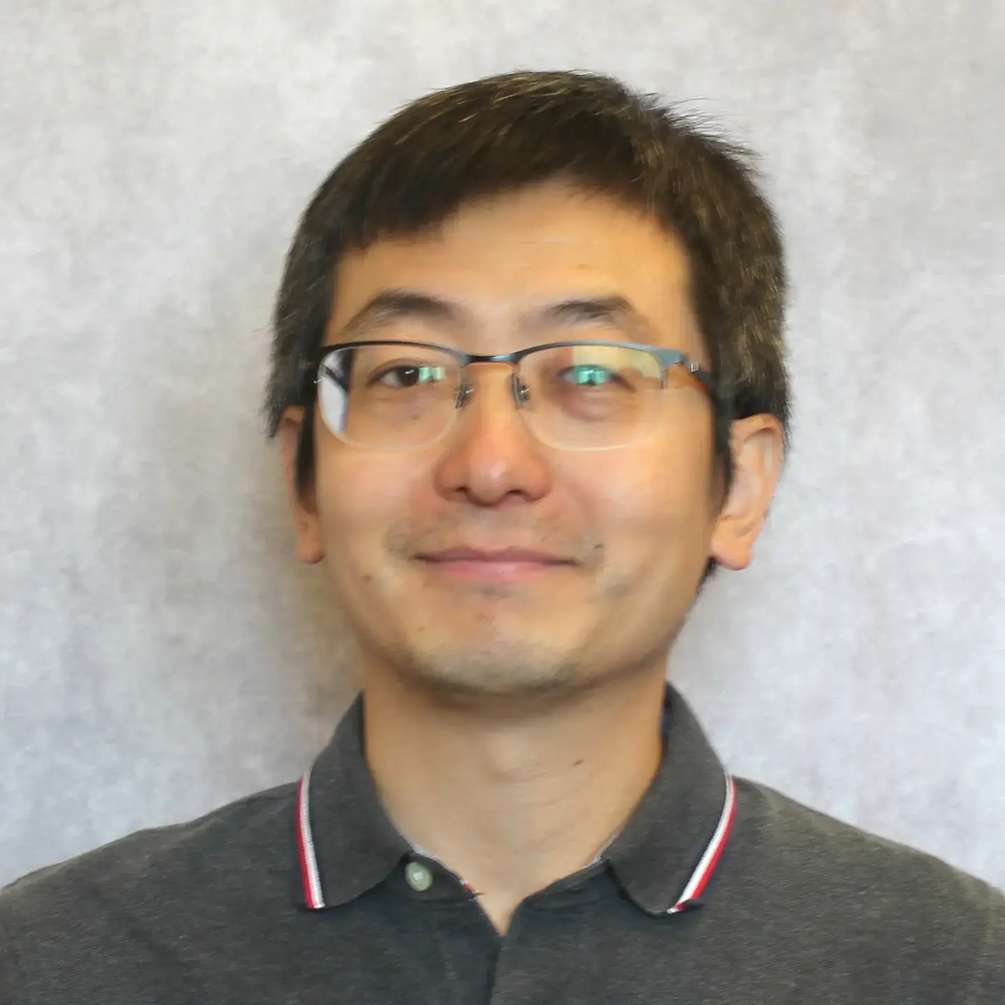 A person with glasses and short black hair, smiling slightly, wearing a gray polo shirt.
