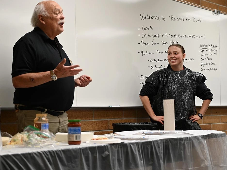 A U of I student wearing a trash bag smiles as a professor speaks in front of a table of supplies to make a pizza in a coding activity