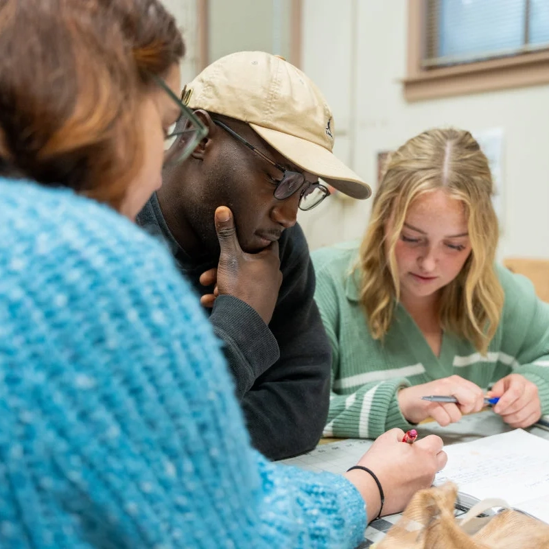 Students ponder their work while seated at a table covered with papers.