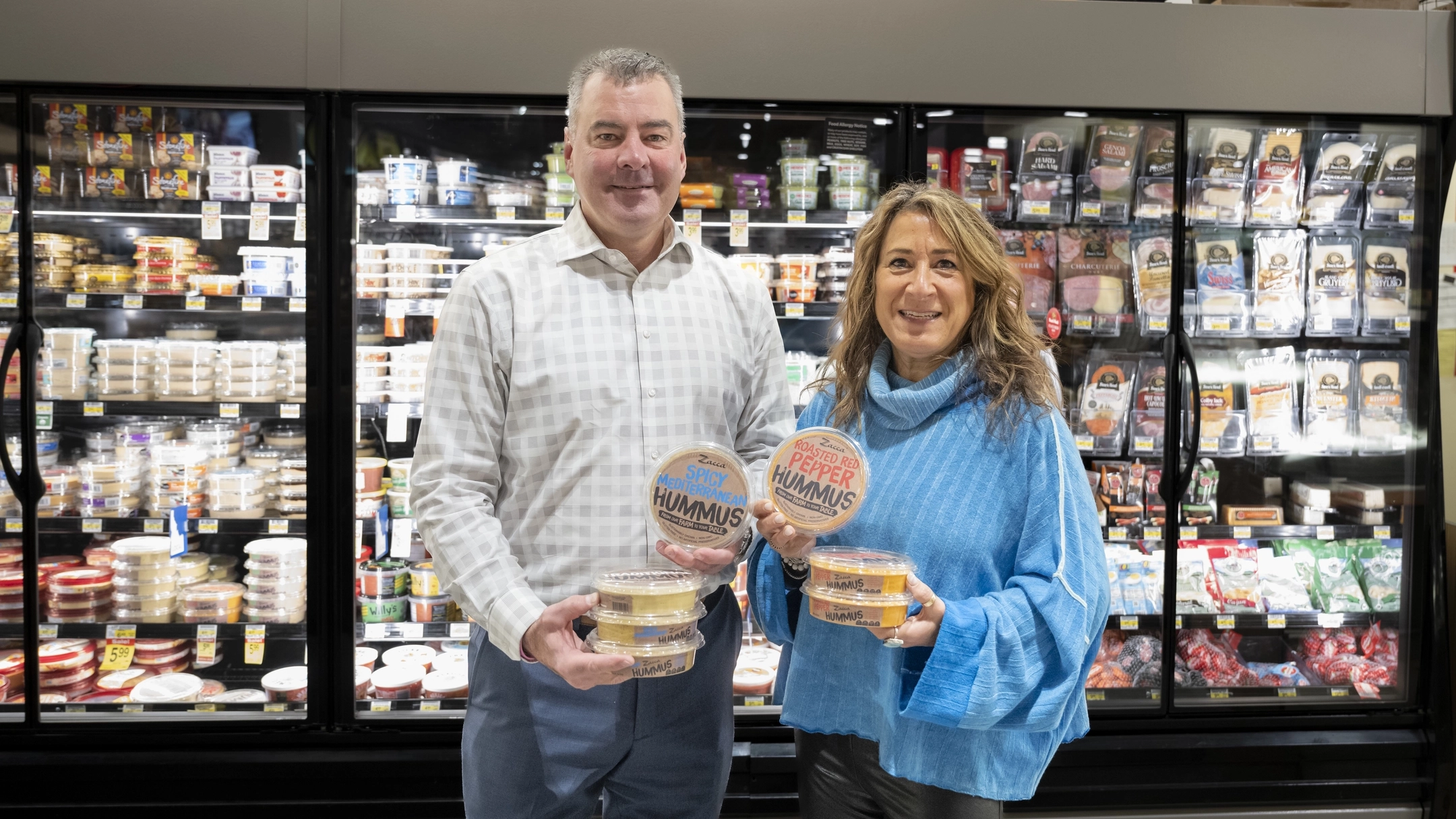 Co-founders of Zacca Hummus Chris, a Vandal Alum, and Janine Zacca Zenner in an Albertsons store in Boise Idaho, holding containers of their Zacca Hummus from a display.