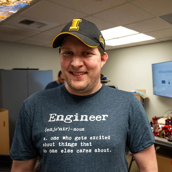 A young man in an Idaho baseball cap smiles for a photo in a robotics classroom