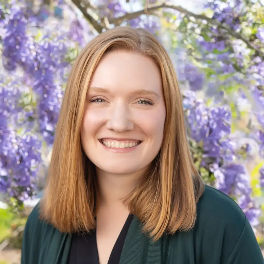 A person smiling outdoors with wisteria flowers in the background.