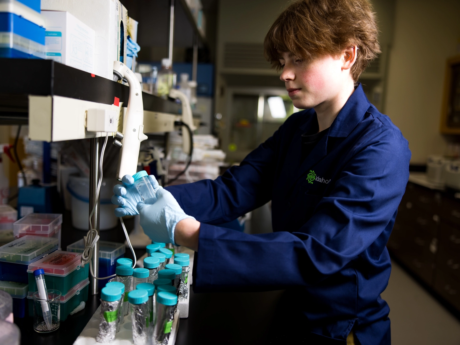 A student wearing a navy-blue lab coat looks at a sample tube in a dim laboratory