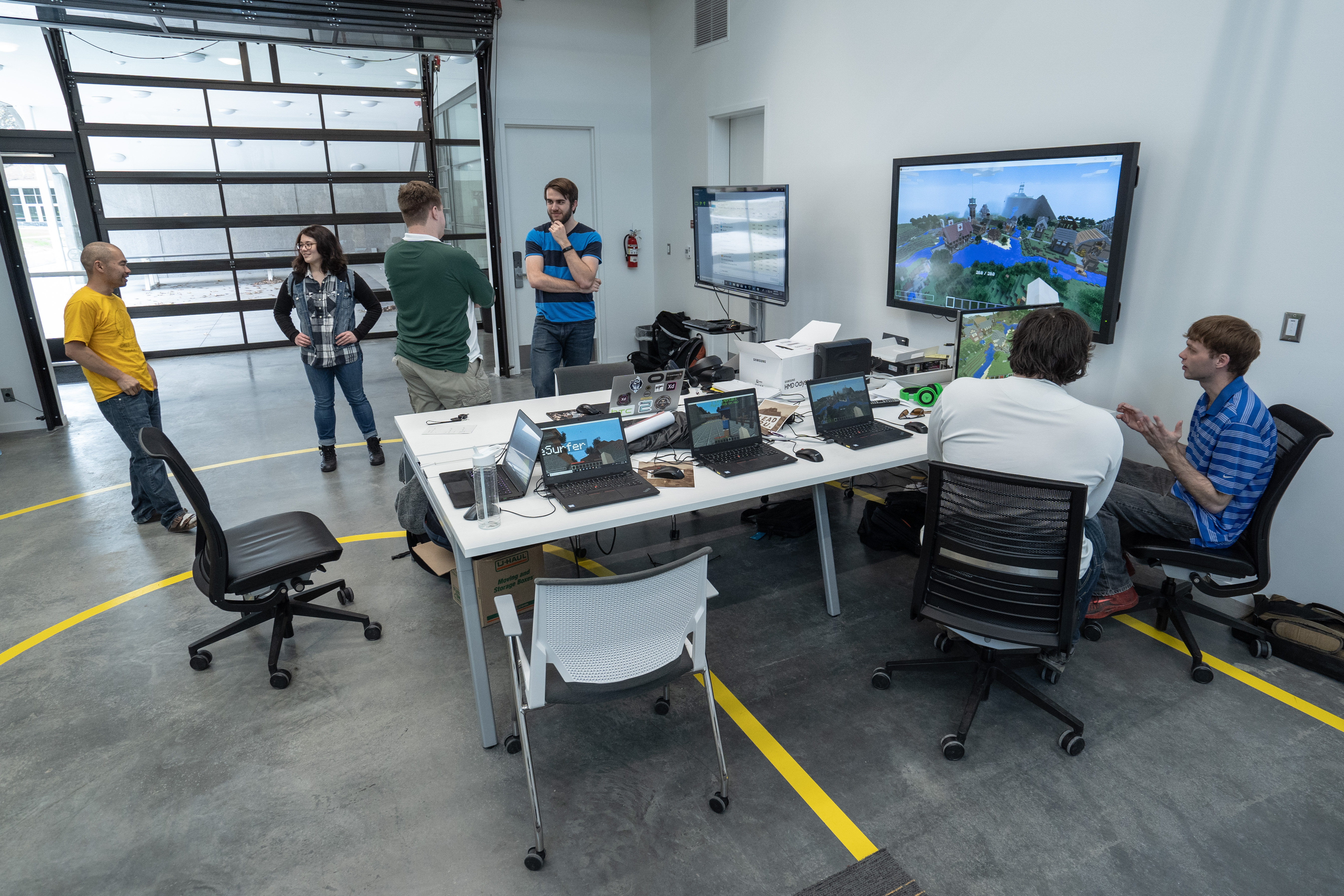 A room with several people interacting. A table with multiple laptops and monitors displays a game setting on one screen.
