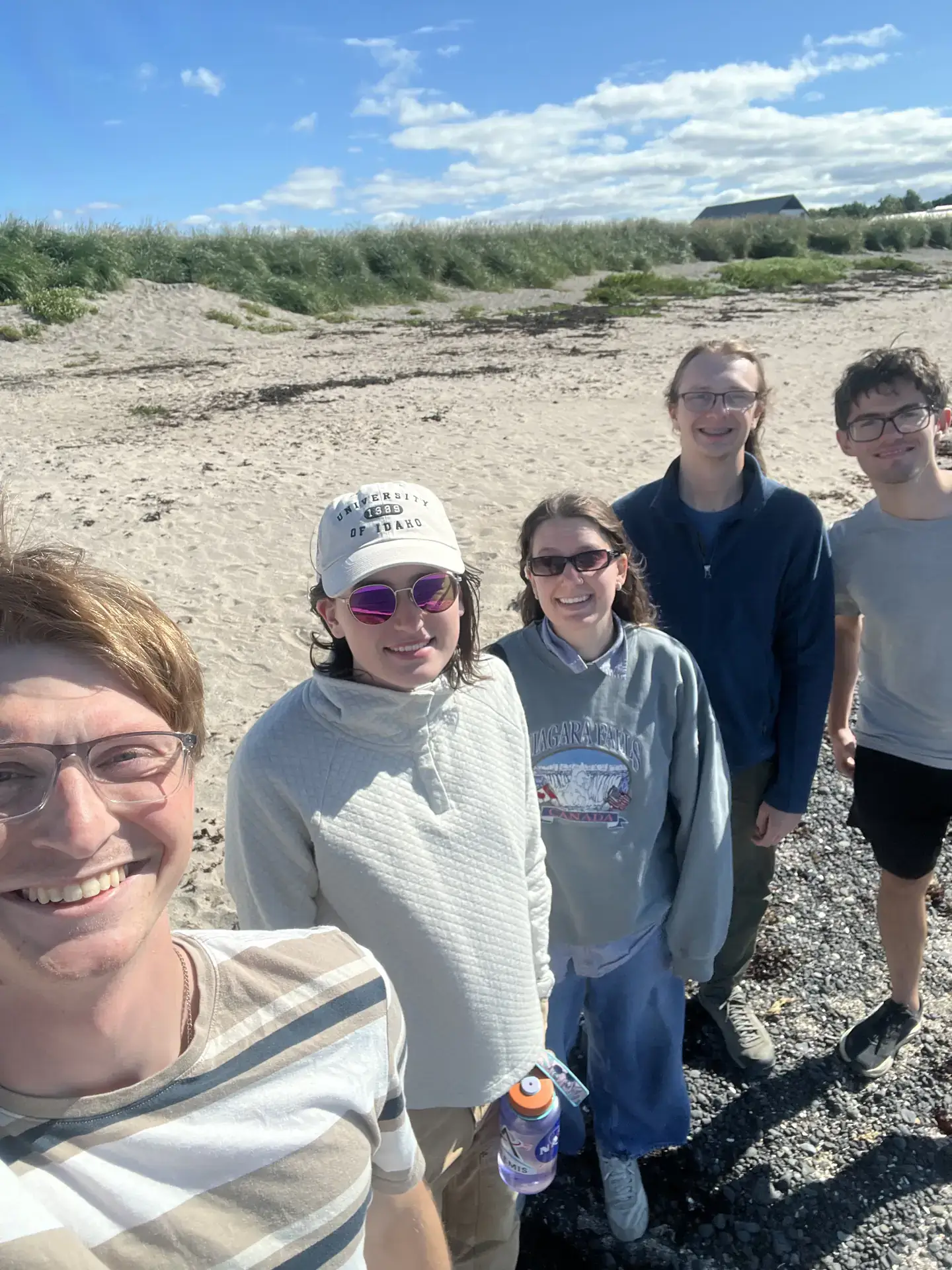 Five young adults smiling for a selfie on a sunny beach with grassy dunes and blue sky behind them.