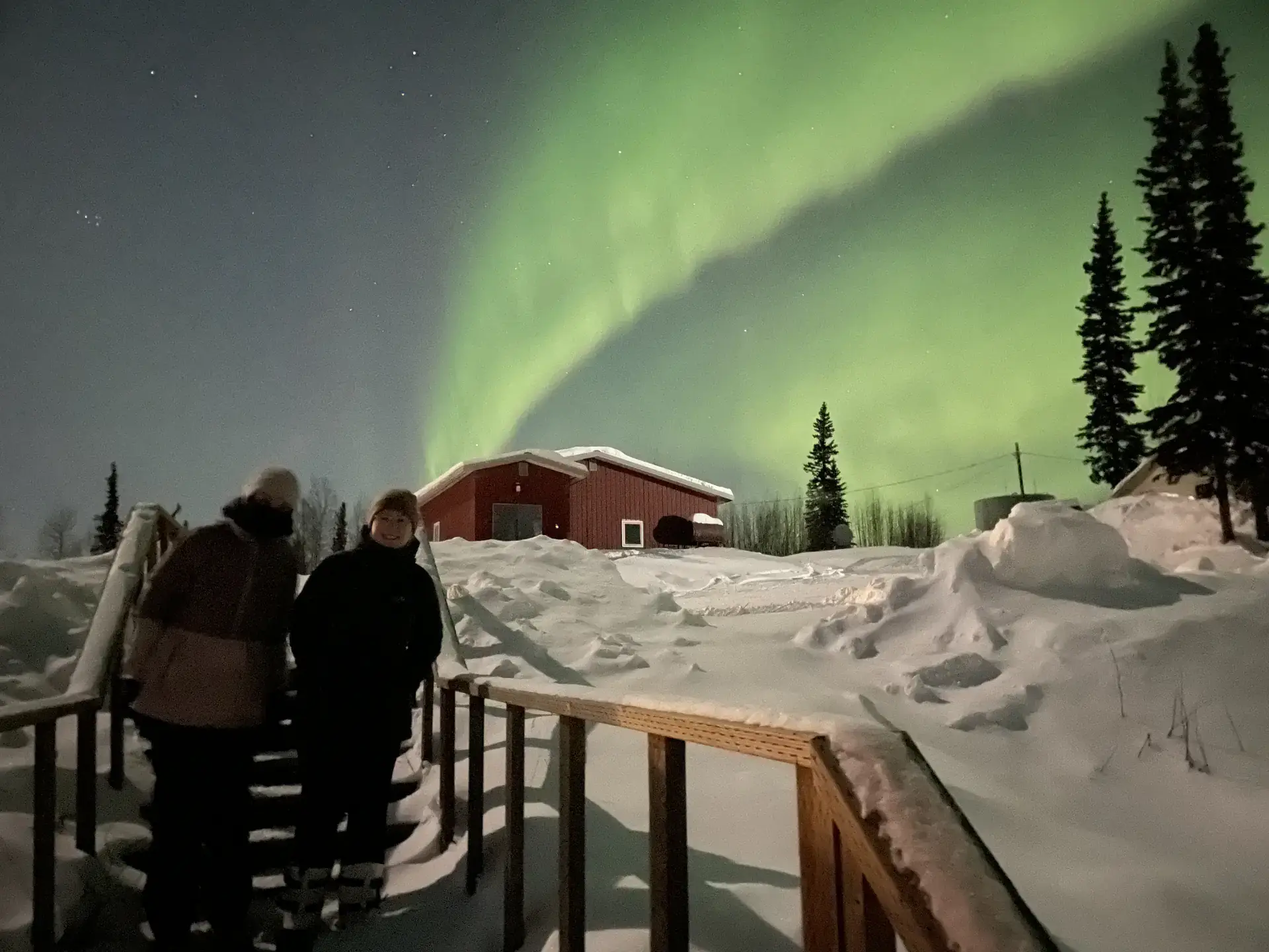 Hannah Westergaard and Alexa Smith stand on a snow-covered walkway. Above them, a turquoise arc of light that bends across the sky.