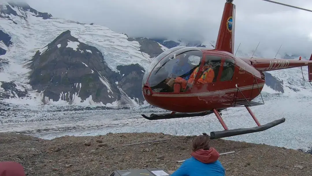 A man kneels near a glacier as a helicopter lands.