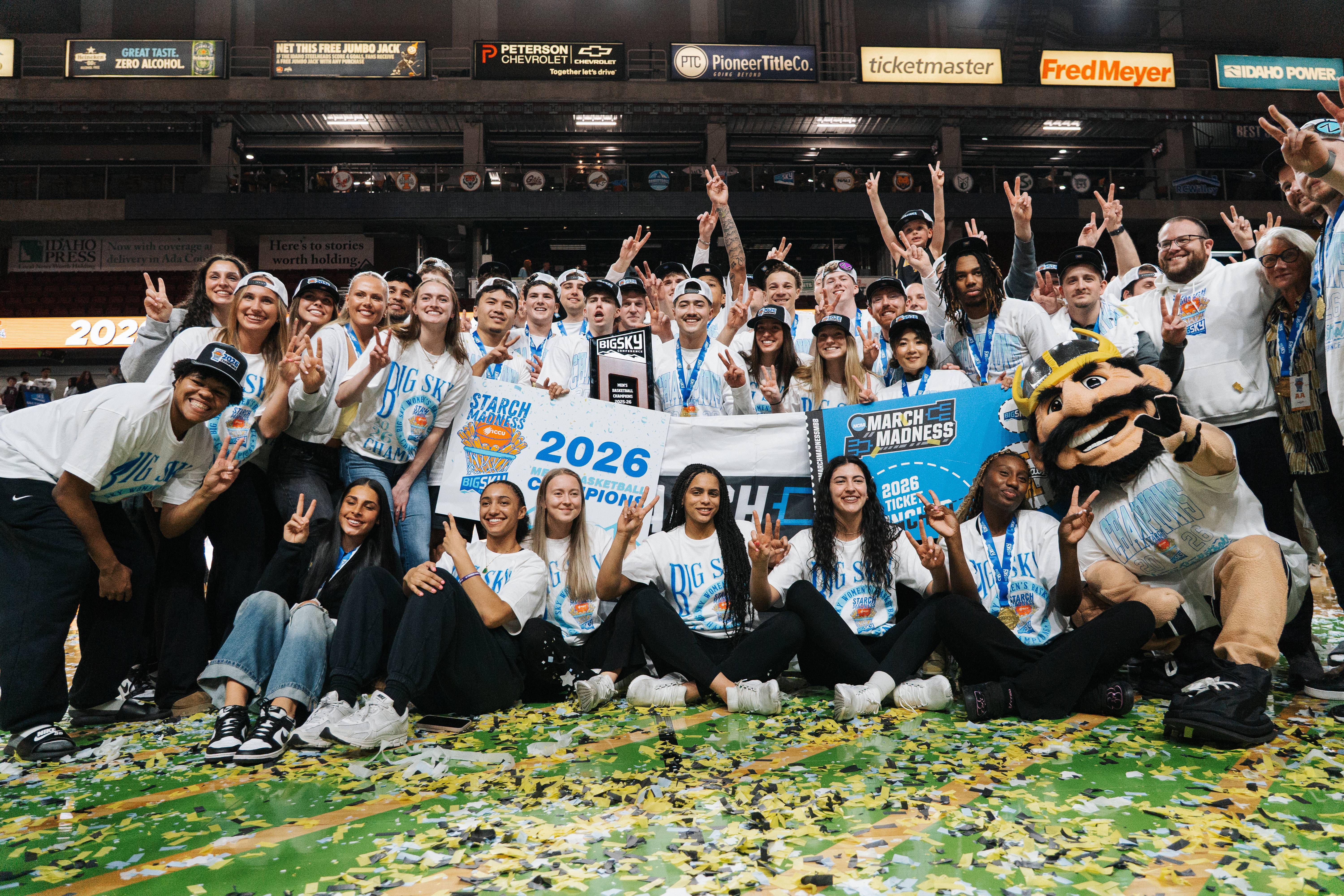 University of Idaho men’s and women's basketball players, students and the Joe Vandal mascot celebrate on a confetti-covered court, holding a “2026 Big Sky Champions” sign and an NCAA March Madness ticket banner while making Vandal hand signs in a packed arena.