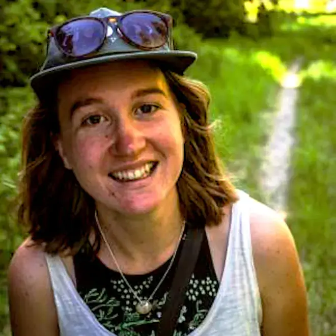 A person smiling outdoors in a sunny field, wearing a hat, sunglasses, a necklace, and a white tank top.