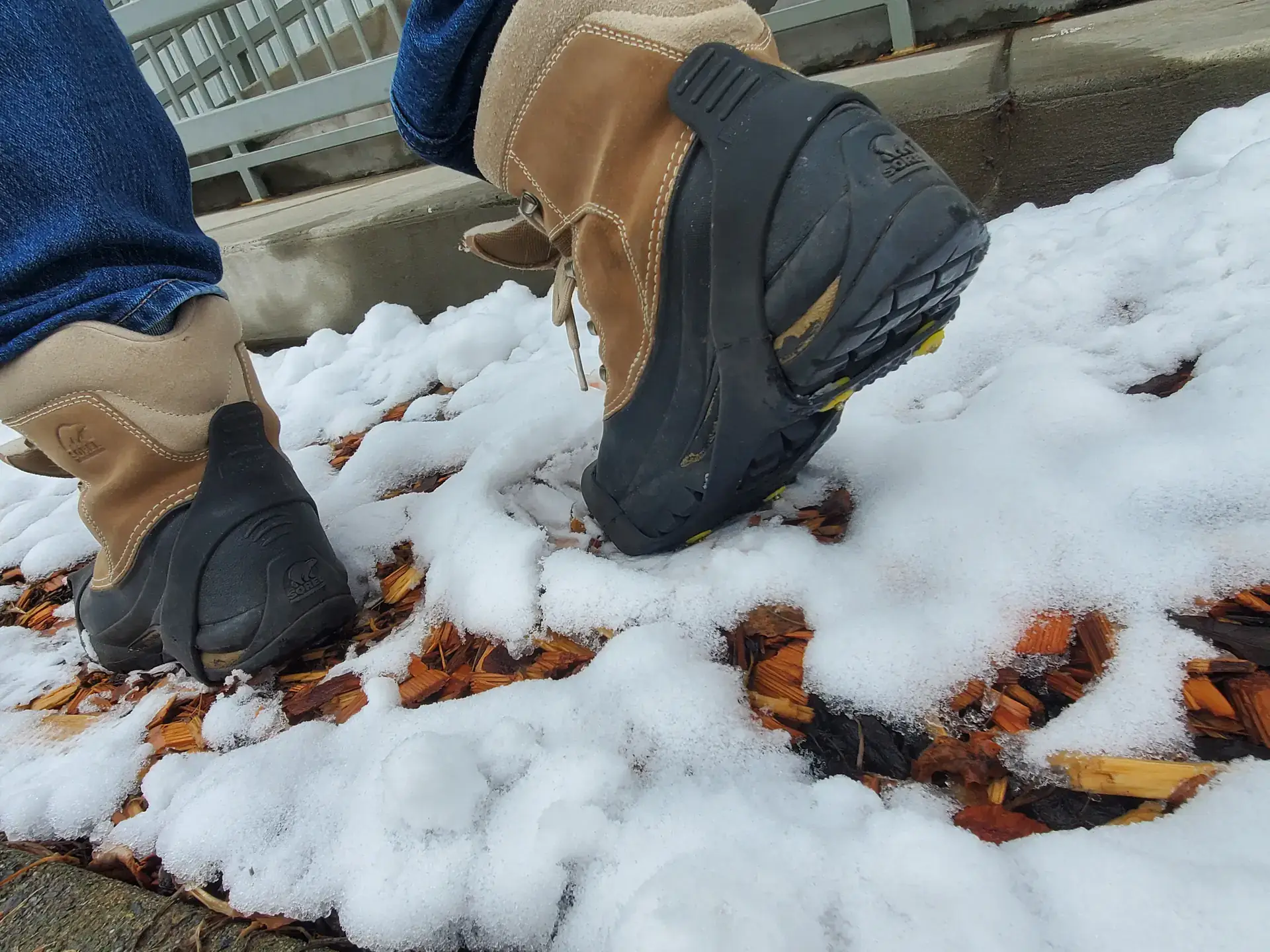 Tan winter boots with traction devices attached, stepping through patchy snow.