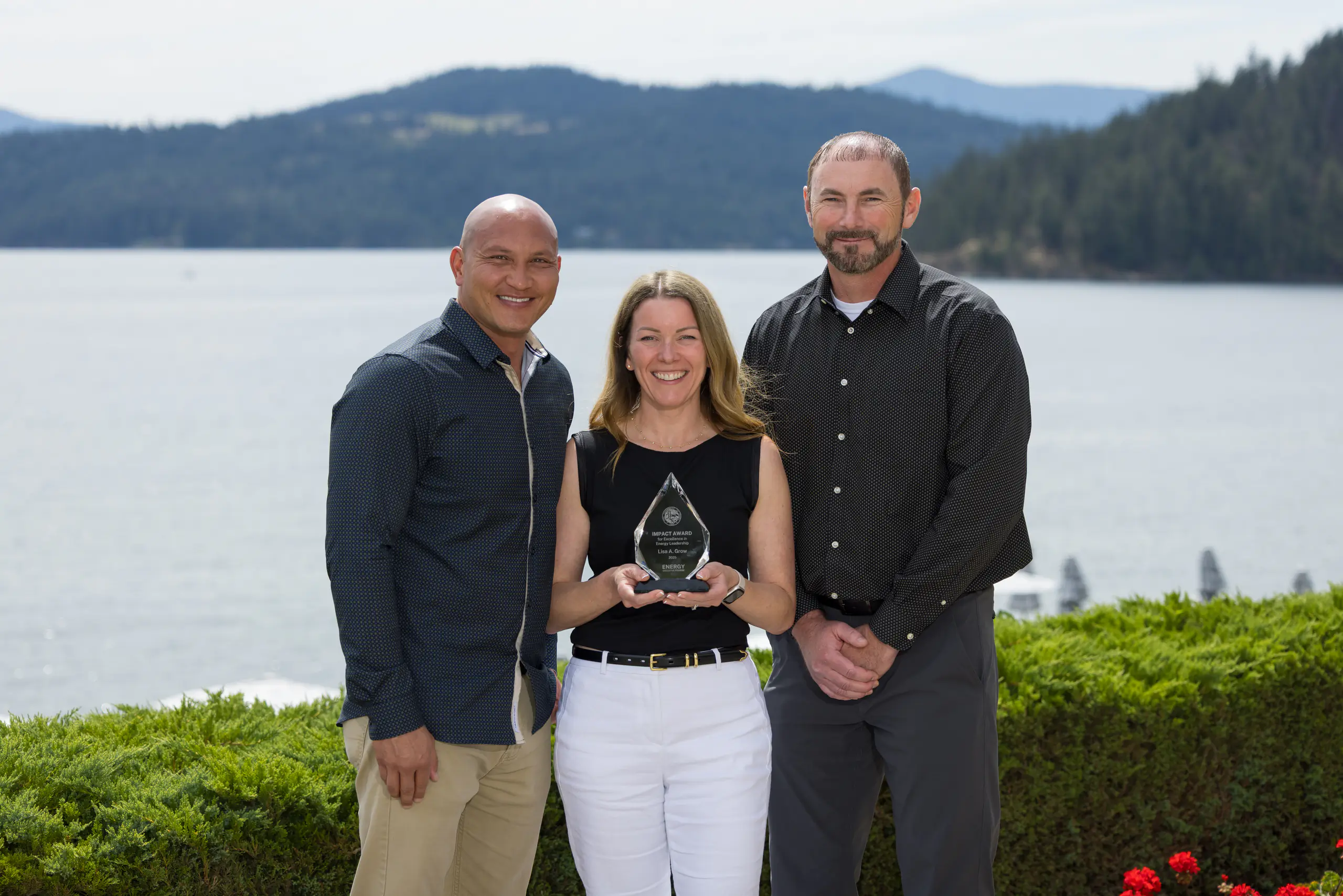 A woman holding an award, with two men at her sides, in front of a lake, wearing business casual attire