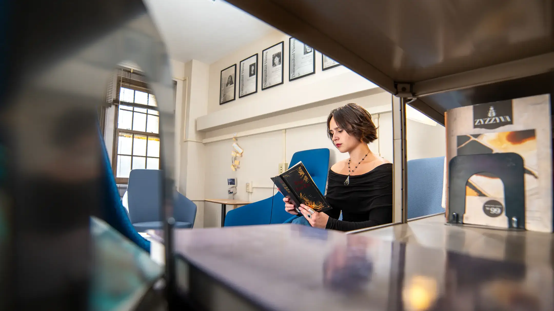 Student seated in a library nook, reading a book between shelves and blue lounge chairs.