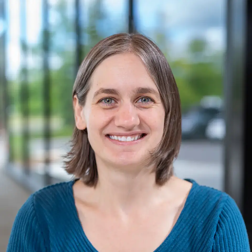 A woman with short brown hair and a blue sweater smiles, with a blurred background that includes windows and greenery.