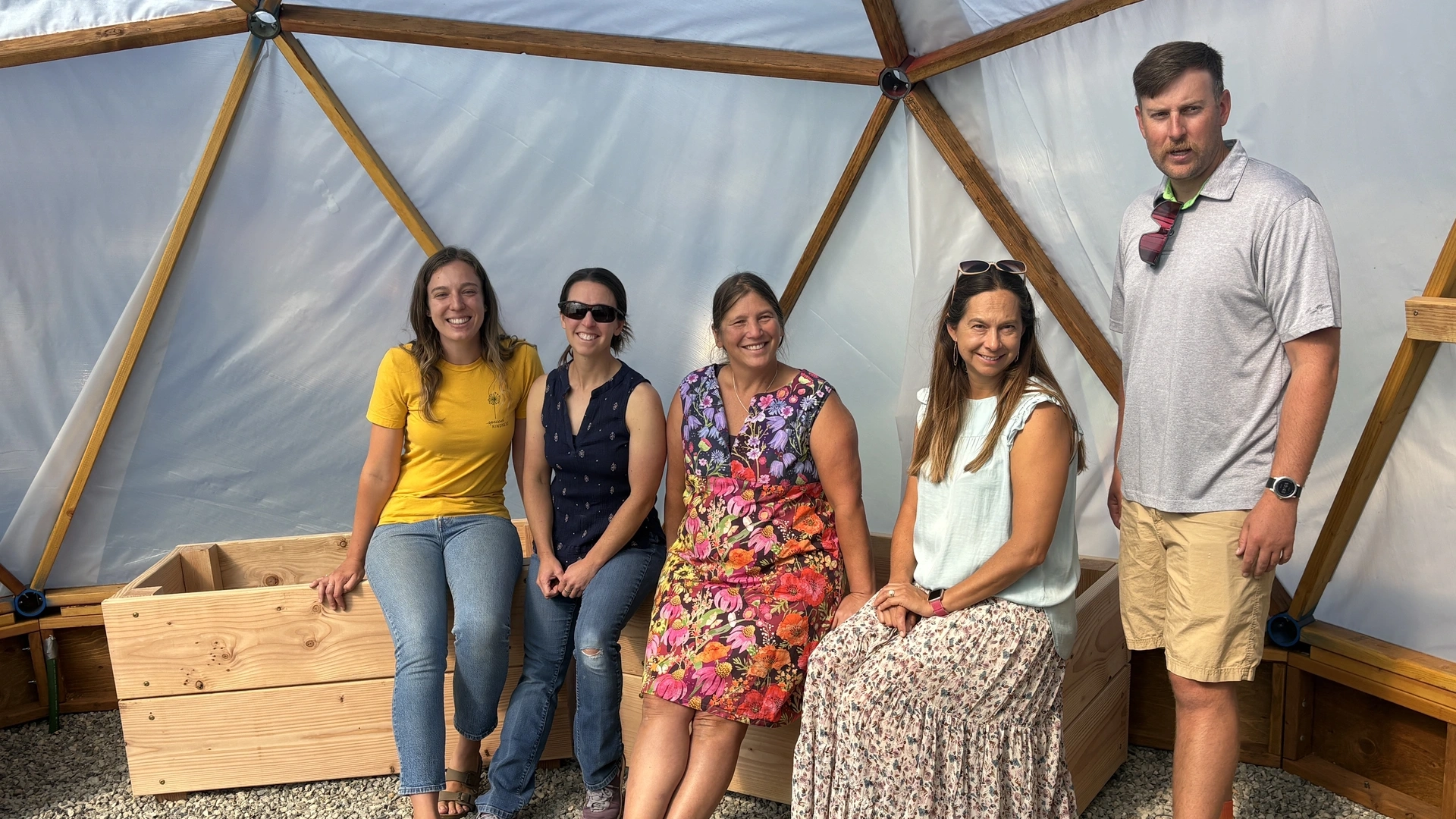 A group pose inside of a geodesic dome greenhouse with a gravel floor and an exterior made of plastic stretched over a frame of boards in triangular patterns.