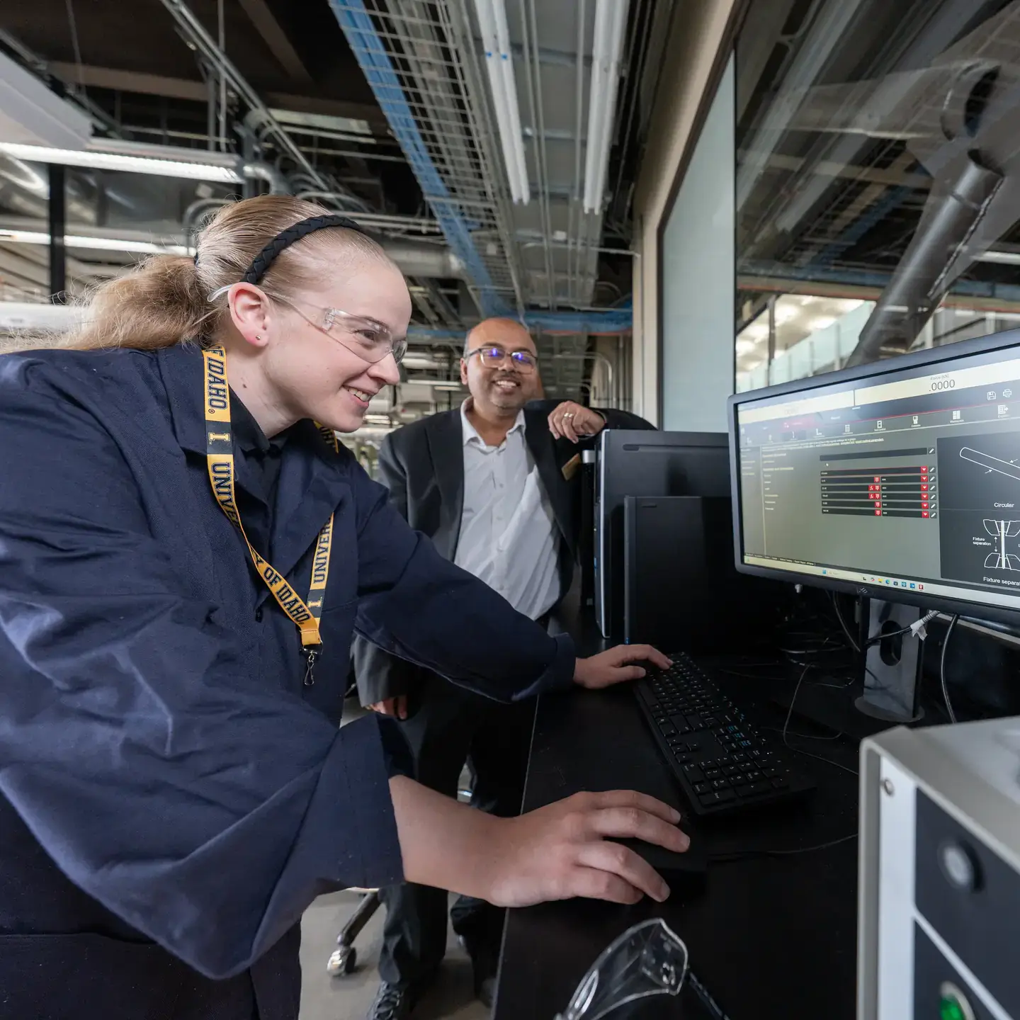 Two individuals in a laboratory setting, one operating a computer system with diagrams displayed on the screen, the other observing.