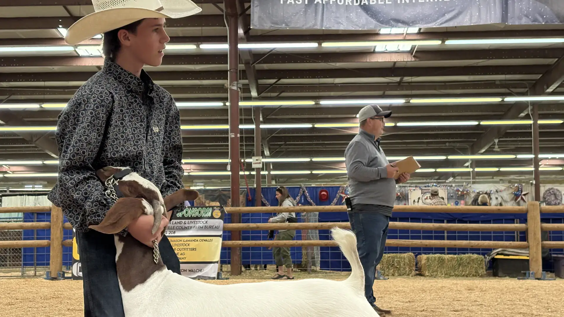 A person in a cowboy hat holds a goat on a leash in a livestock arena with visible banners and spectators in the background.