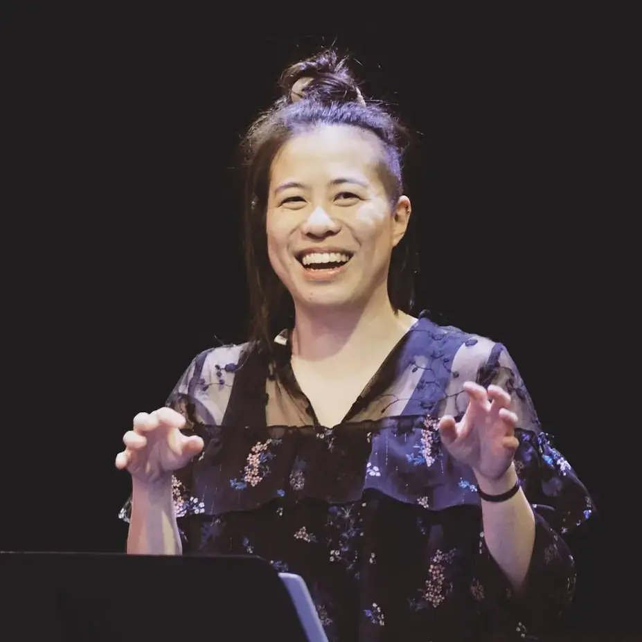 A person smiling on stage, wearing a sheer black floral top, with a microphone stand in front of them and a dark background.