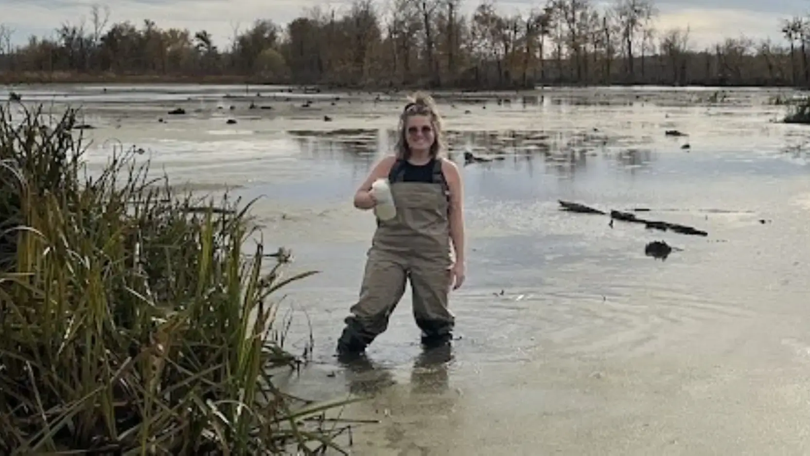 Woman wearing waders stands in a pond holding a jar of water.