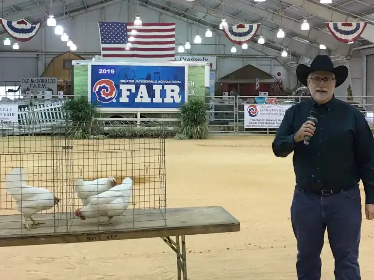 A man wearing a cowboy hat speaks into a microphone in a barn-like setting with American flags and decorative banners. White chickens are in cages resting on a table in the foreground.