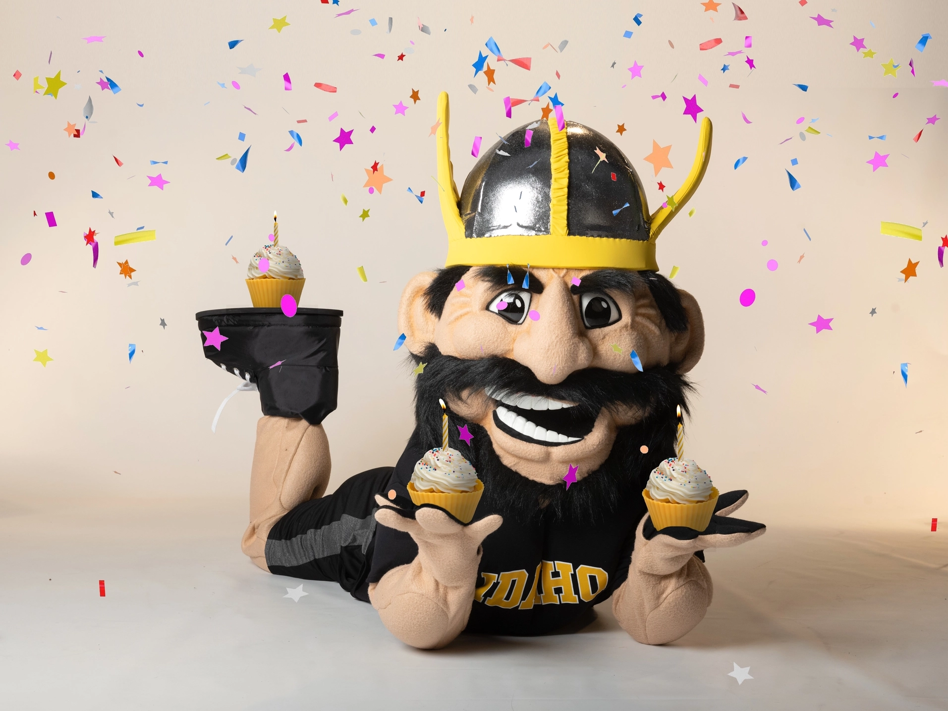 U of I mascot Joe Vandal laying on his stomach holding cupcakes in each hand while a cupcake sits on his foot in the air.