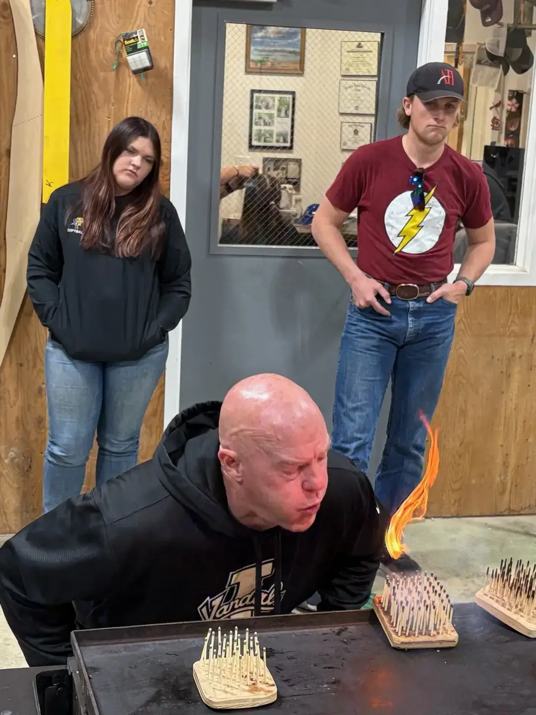 A man leans over a small fire with nails organized on a board, watched by three observers in a workshop setting.