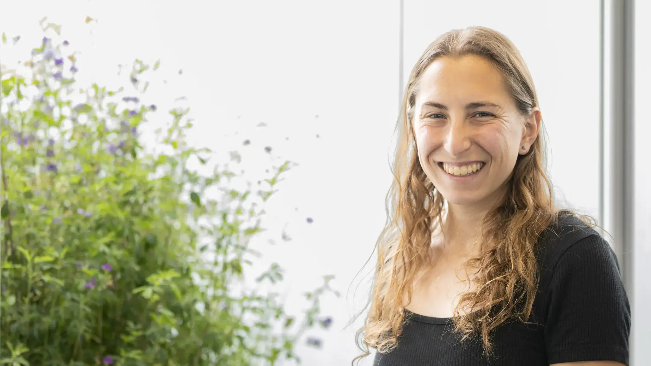 Smiling woman with long wavy hair in a black top, standing indoors beside green plants.