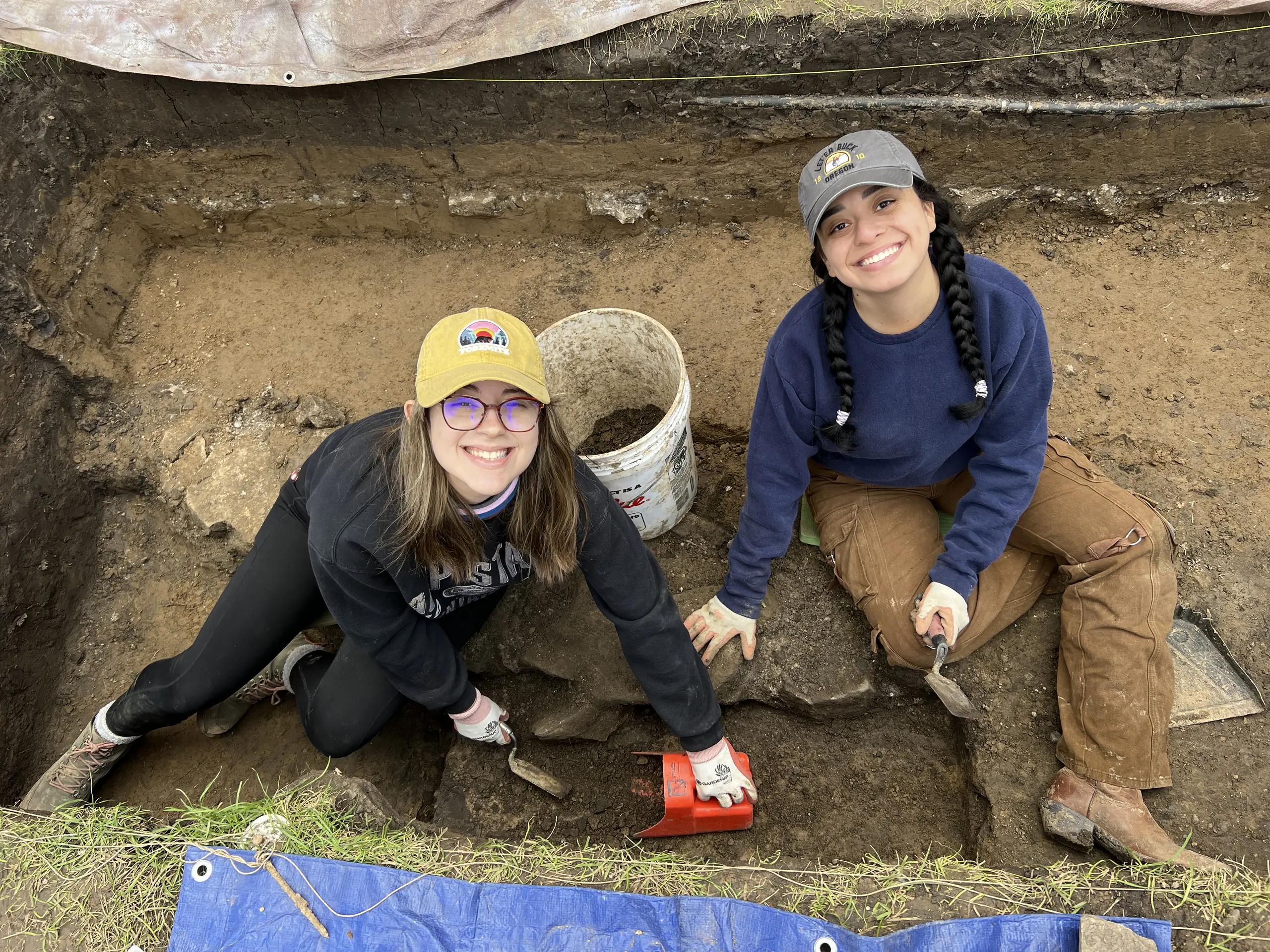 Two archaeology students sit in a rectangular dirt hole with shovels and buckets