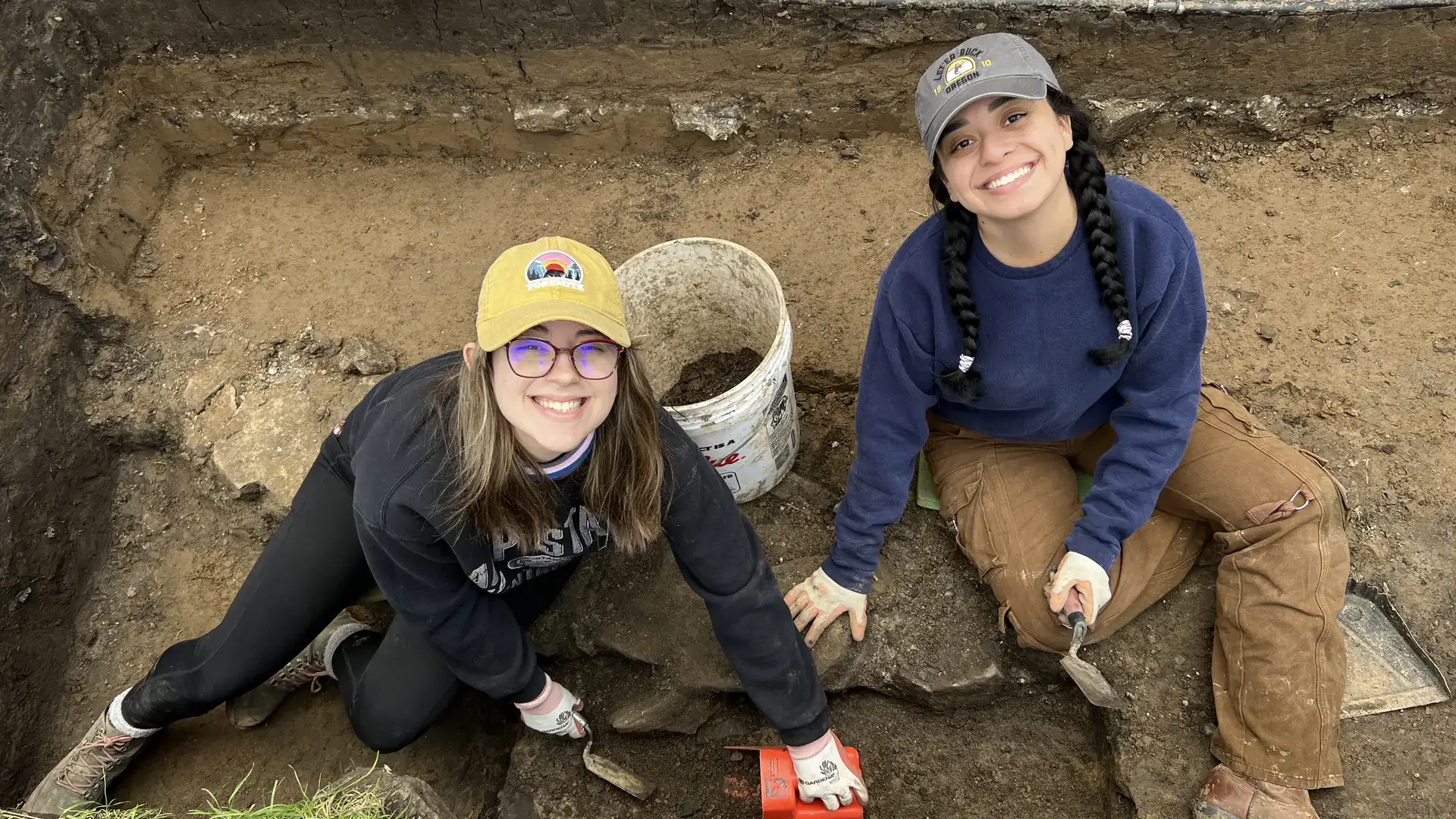 Two archaeology students sit in a rectangular dirt hole with shovels and buckets