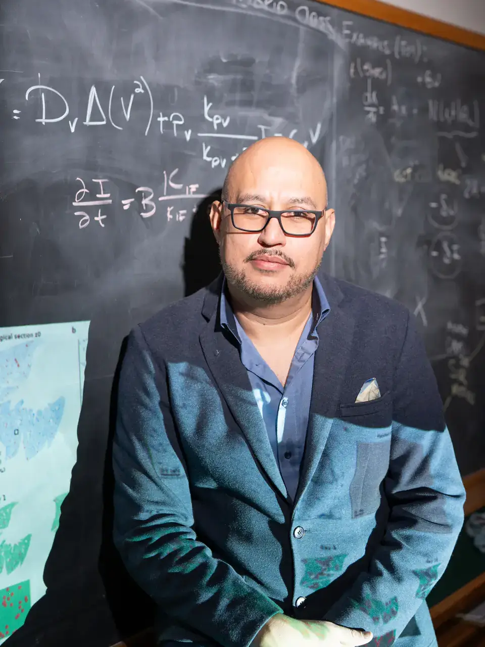 Man in glasses and blazer stands before a chalkboard covered in equations, with scientific images projected on him.