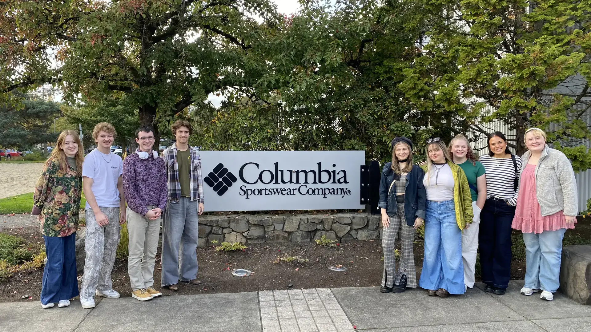 A group of people posing next to a Columbia Sportswear Company sign surrounded by trees on a pathway outdoors.