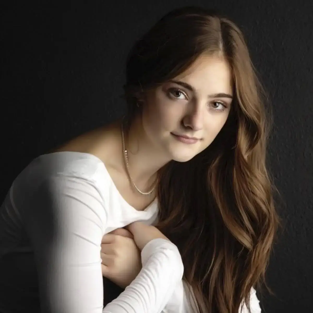 A person with long brown hair wearing a white top, sitting against a dark background.