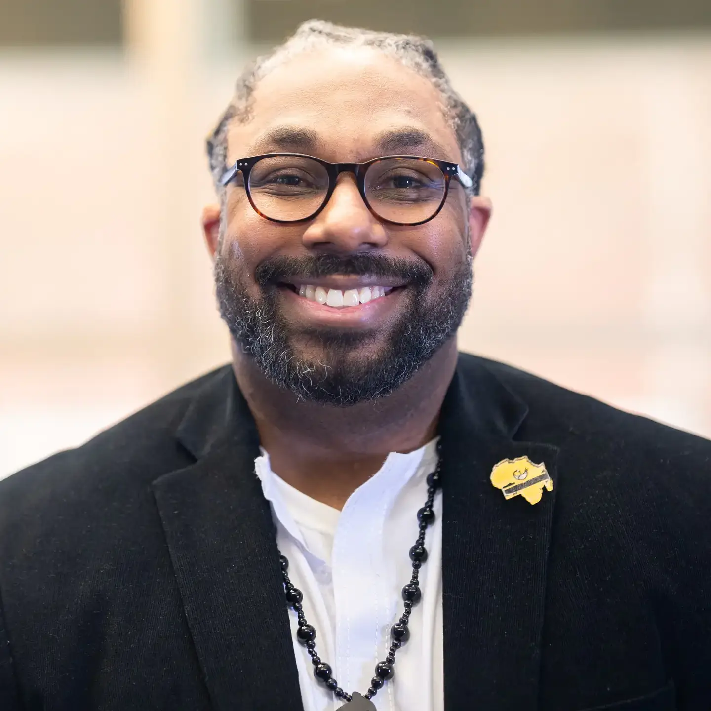 A person wearing glasses, a black blazer, and a white shirt. They are smiling and have a pin attached to their blazer.