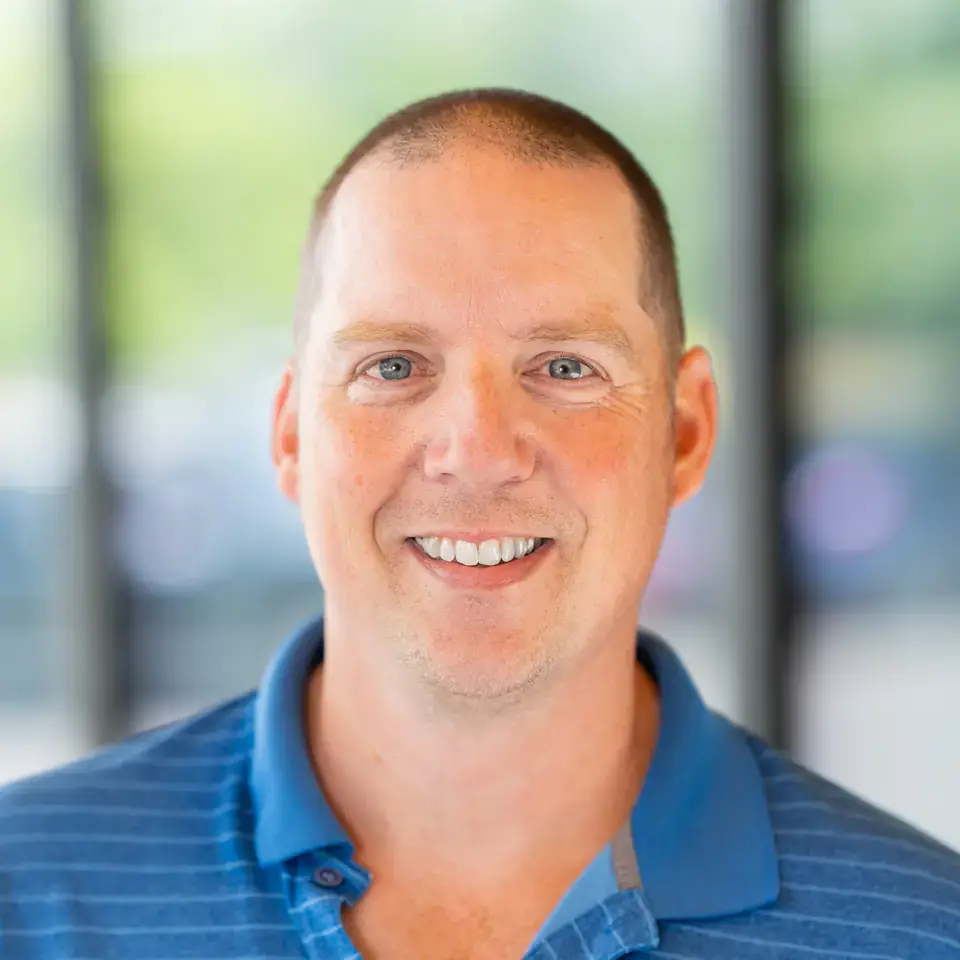 A man in a blue striped polo shirt smiles at the camera with a blurred outdoor background.