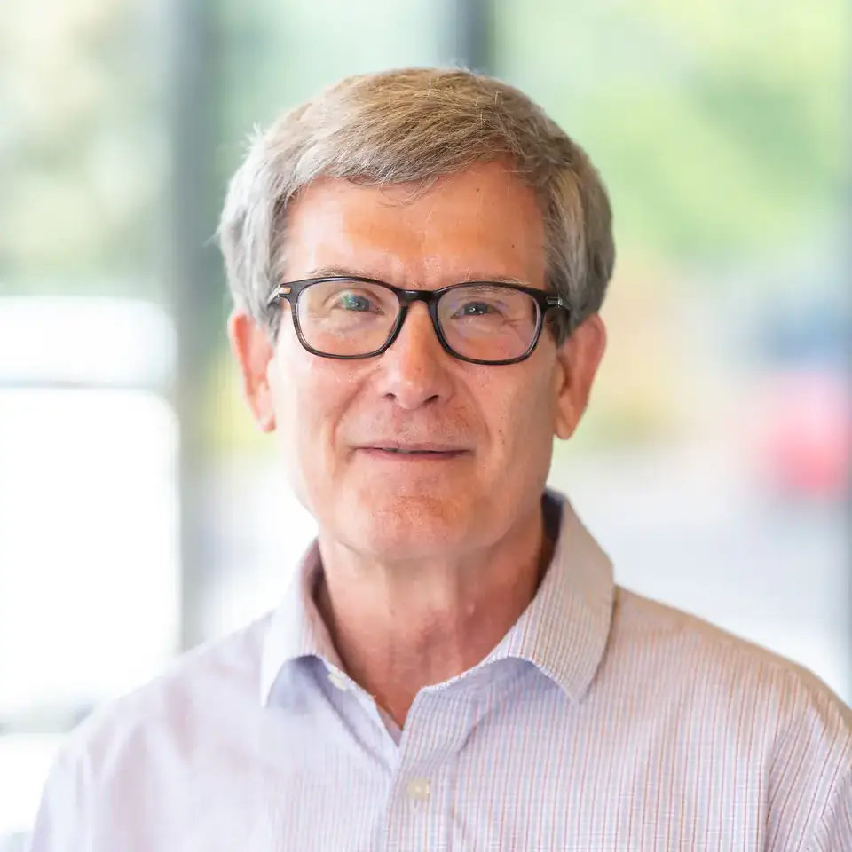 Portrait of an individual wearing glasses and a checkered shirt, with a blurred background featuring a window and outdoor greenery.