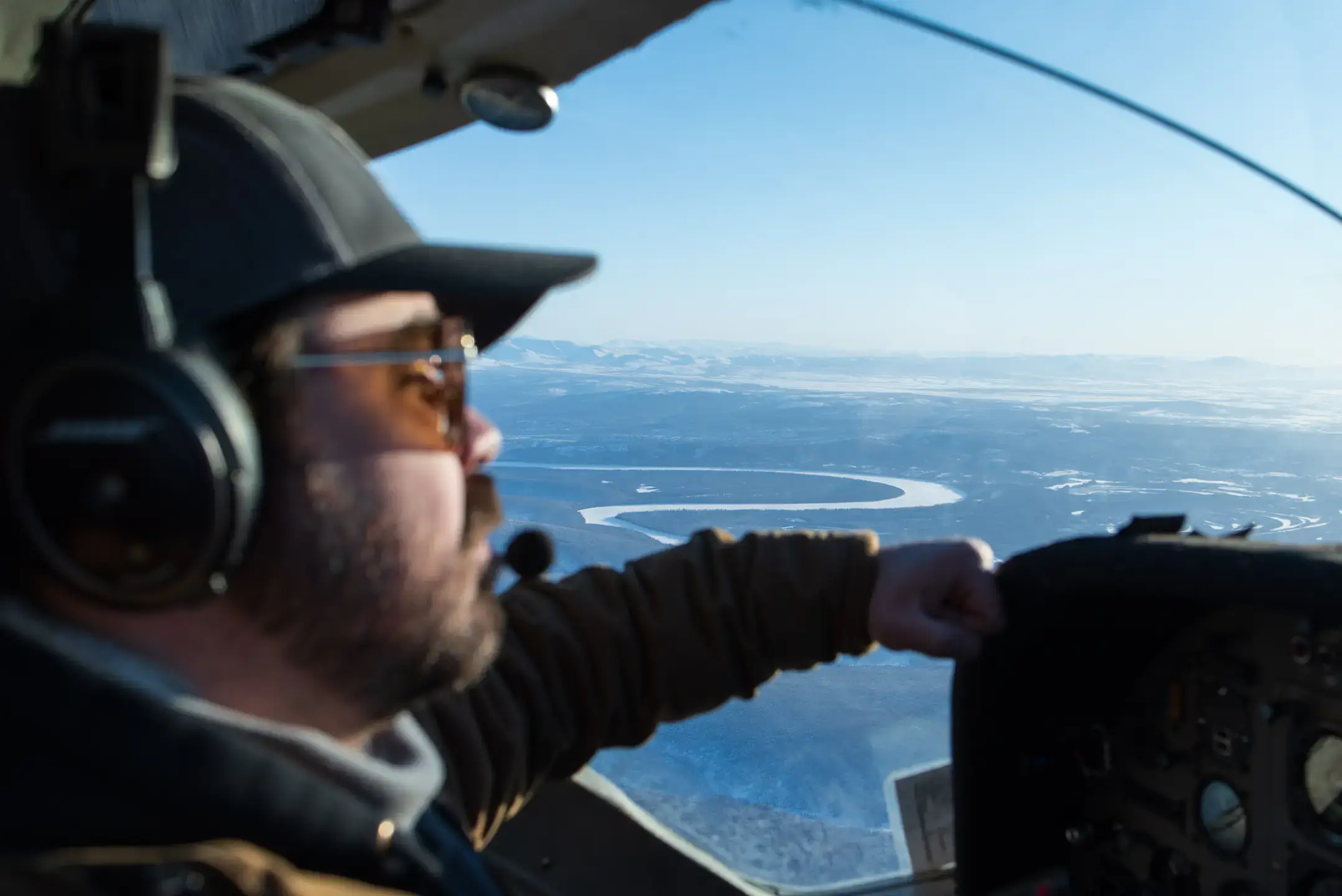 Over the right shoulder of an aircraft pilot, the Alaskan frontier extends toward the horizon: a winding river, mountains and trees are far below.