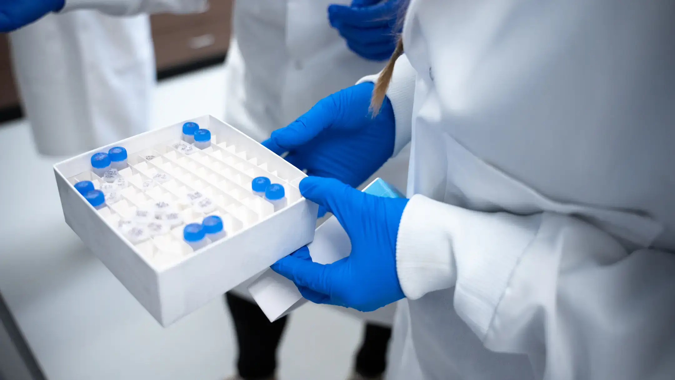 A person in a white lab coat with blue, rubber gloves holds a box of test tubes.