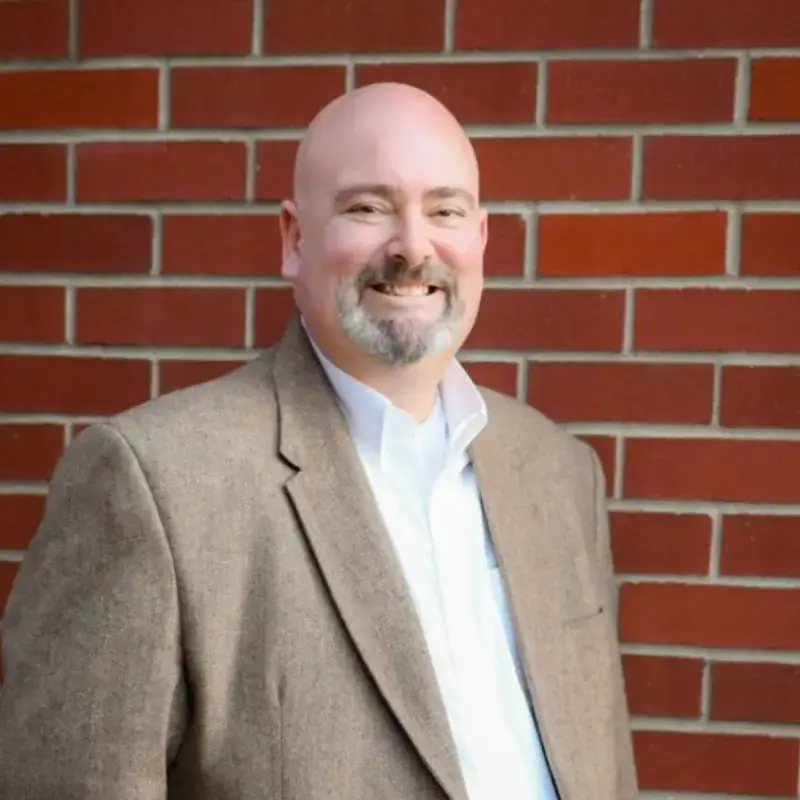 A person wearing a suit jacket and shirt smiles in front of a red brick wall.
