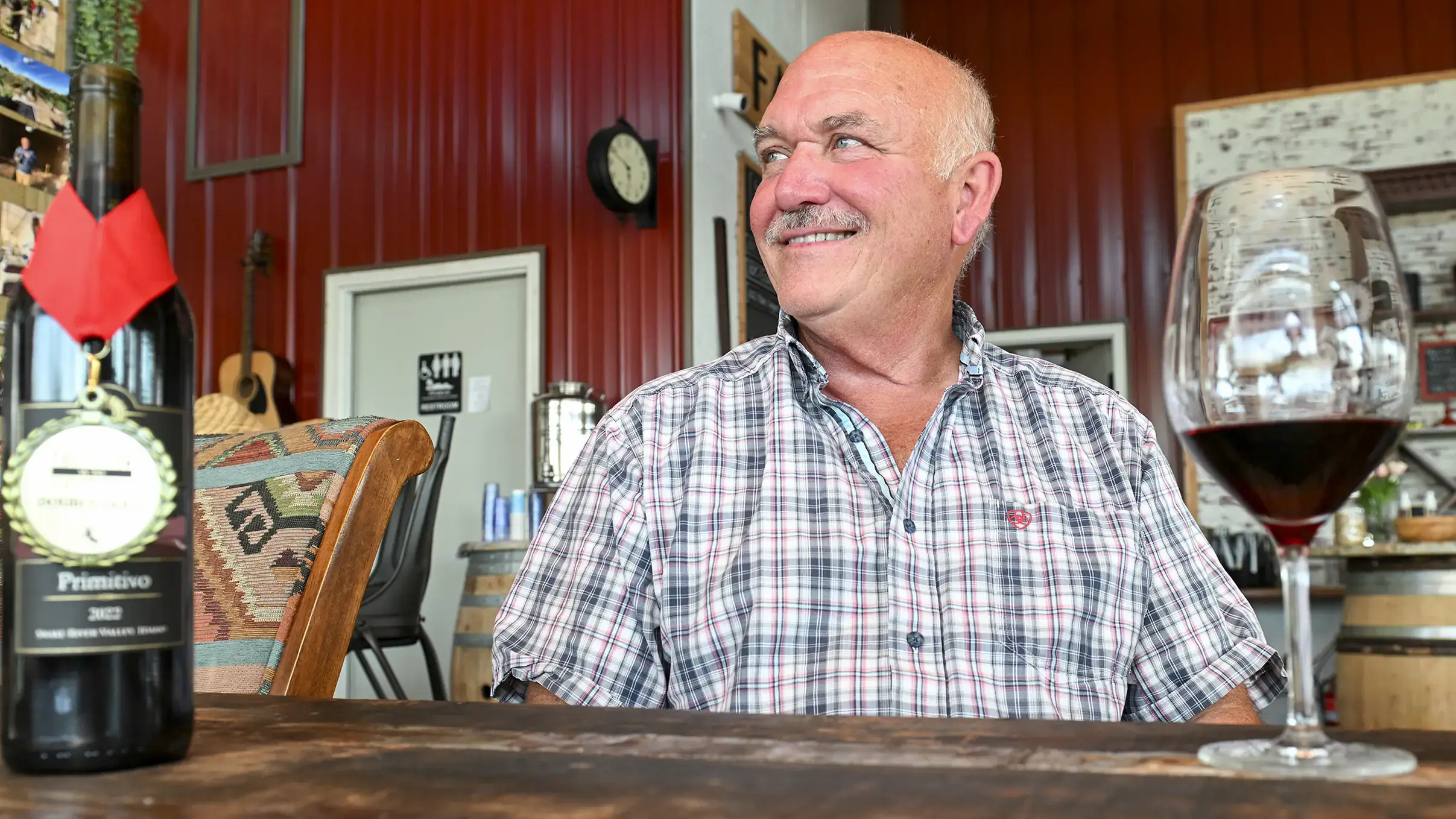 A person sits smiling in a wine tasting room with a glass of red wine and a bottle labeled Primitivo 2021 on a wooden table.