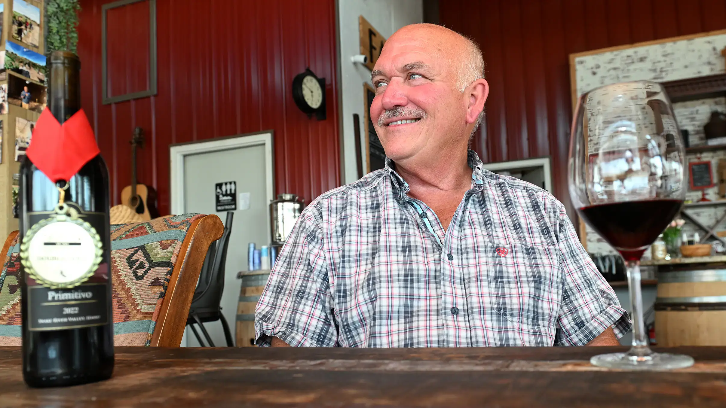 A man sits at a wooden table in a winery, smiling. A bottle of wine and a glass of red wine are in the foreground.