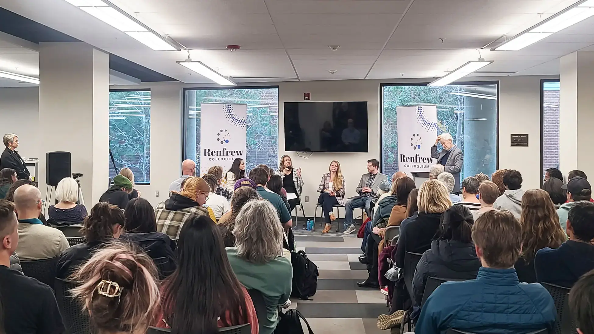 An audience sits facing a panel of speakers in a modern seminar room with windows, banners, and overhead lighting.