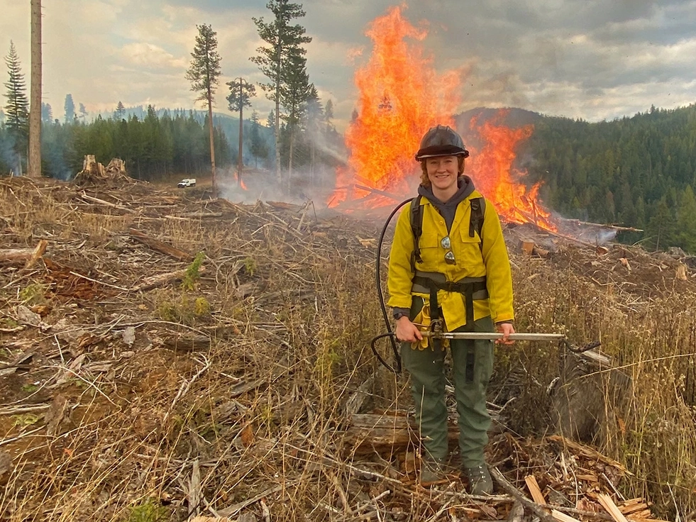 A woman wearing wildland fire gear stands near a burning slash fire