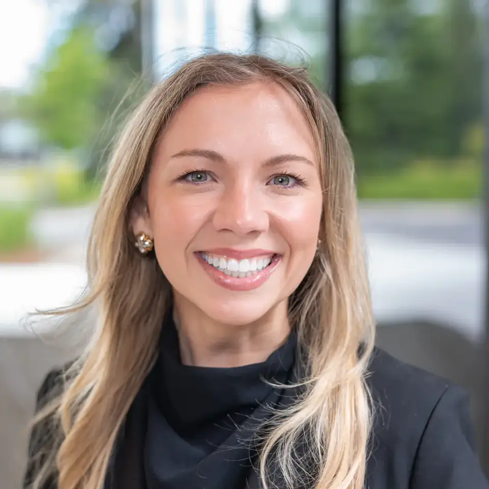 A woman smiling against a blurred outdoor background, wearing a black blazer and long hair flowing over her shoulders.