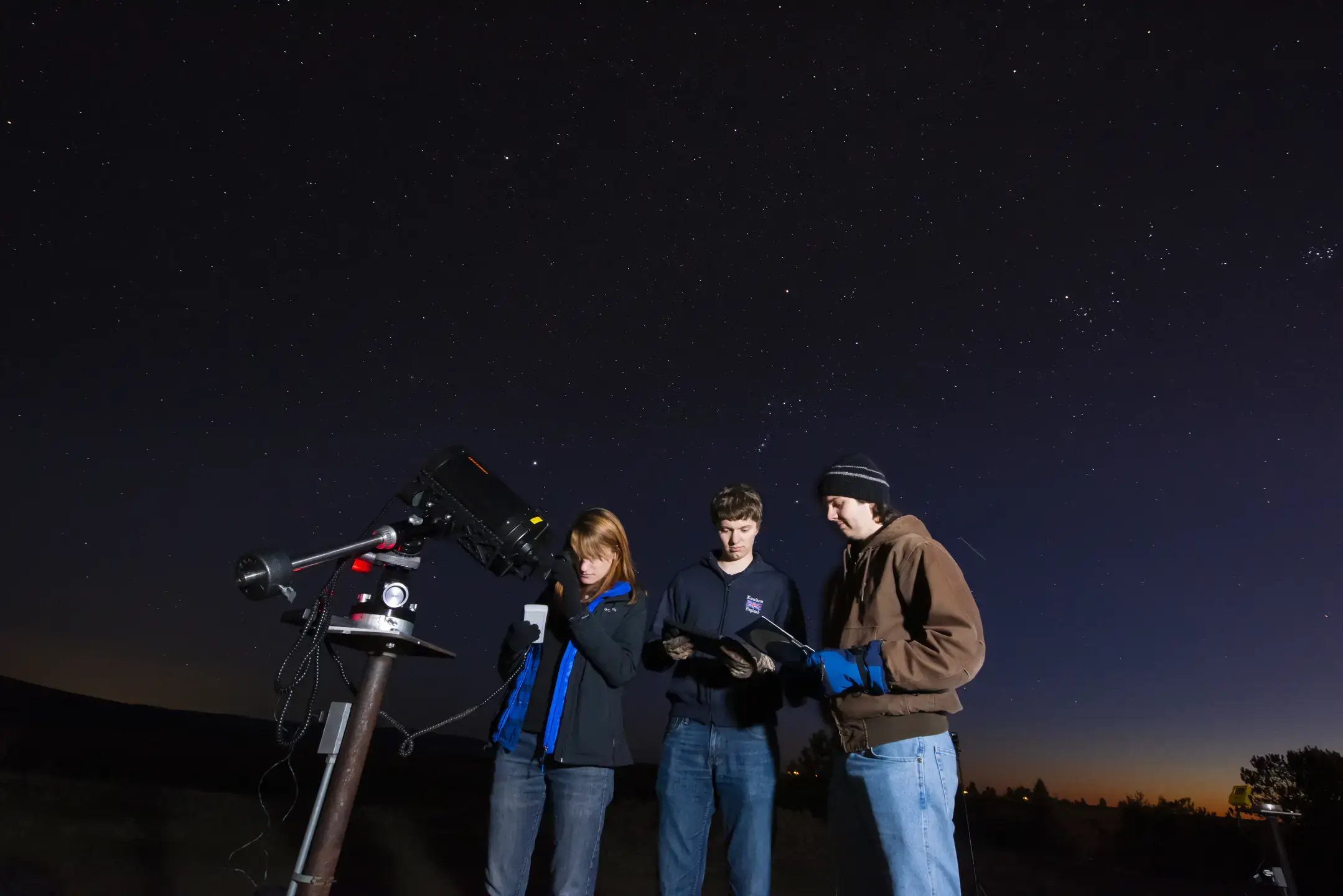 Three people work with a telescope at night under a clear, star-filled sky.