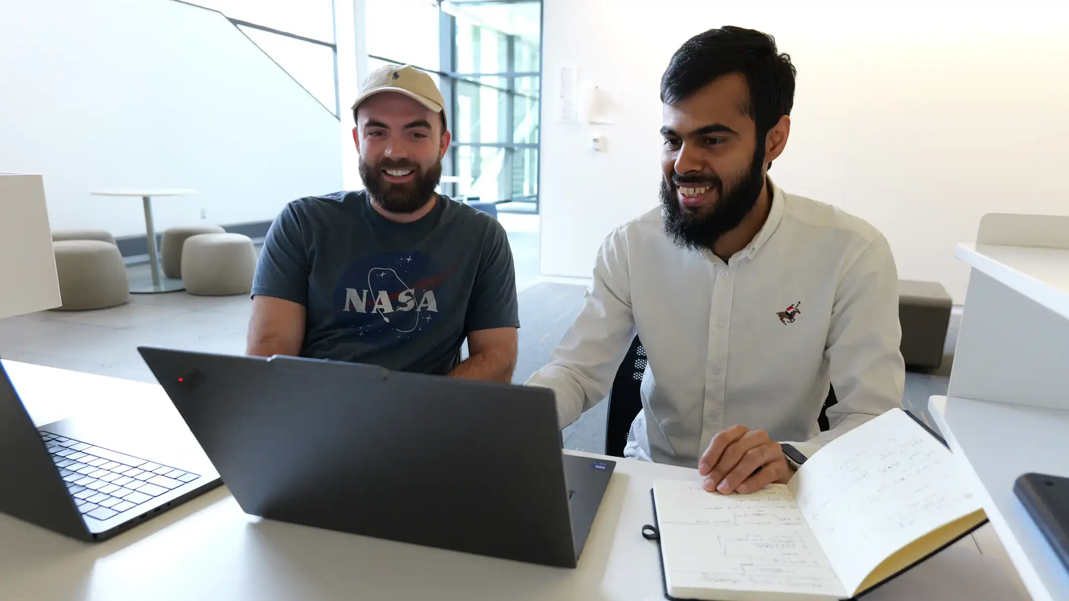 Two men collaborate at laptops with a notebook open on a desk in a bright office.