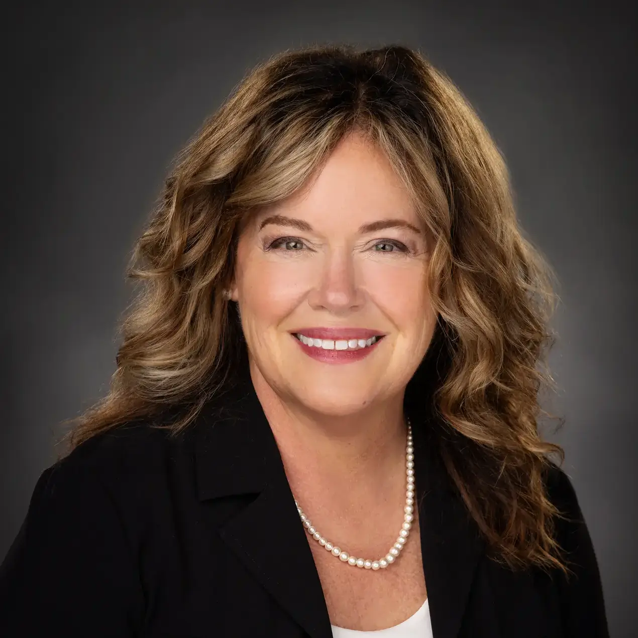 Professional portrait of a woman with curly hair, wearing a black blazer and pearl necklace, smiling against a dark background.