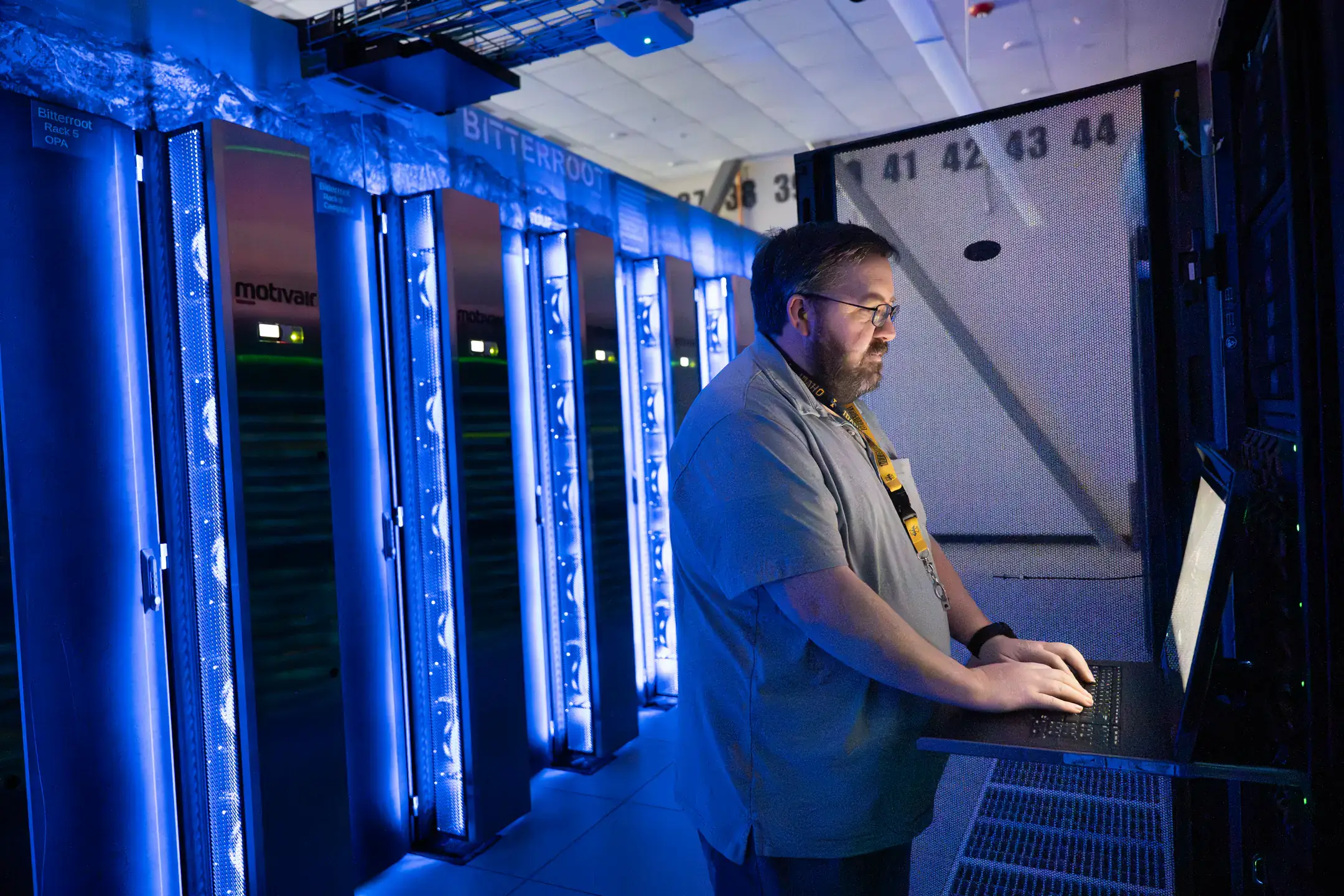 A man works on a laptop in a server room with blue-lit racks labeled 'Bitterroot'.
