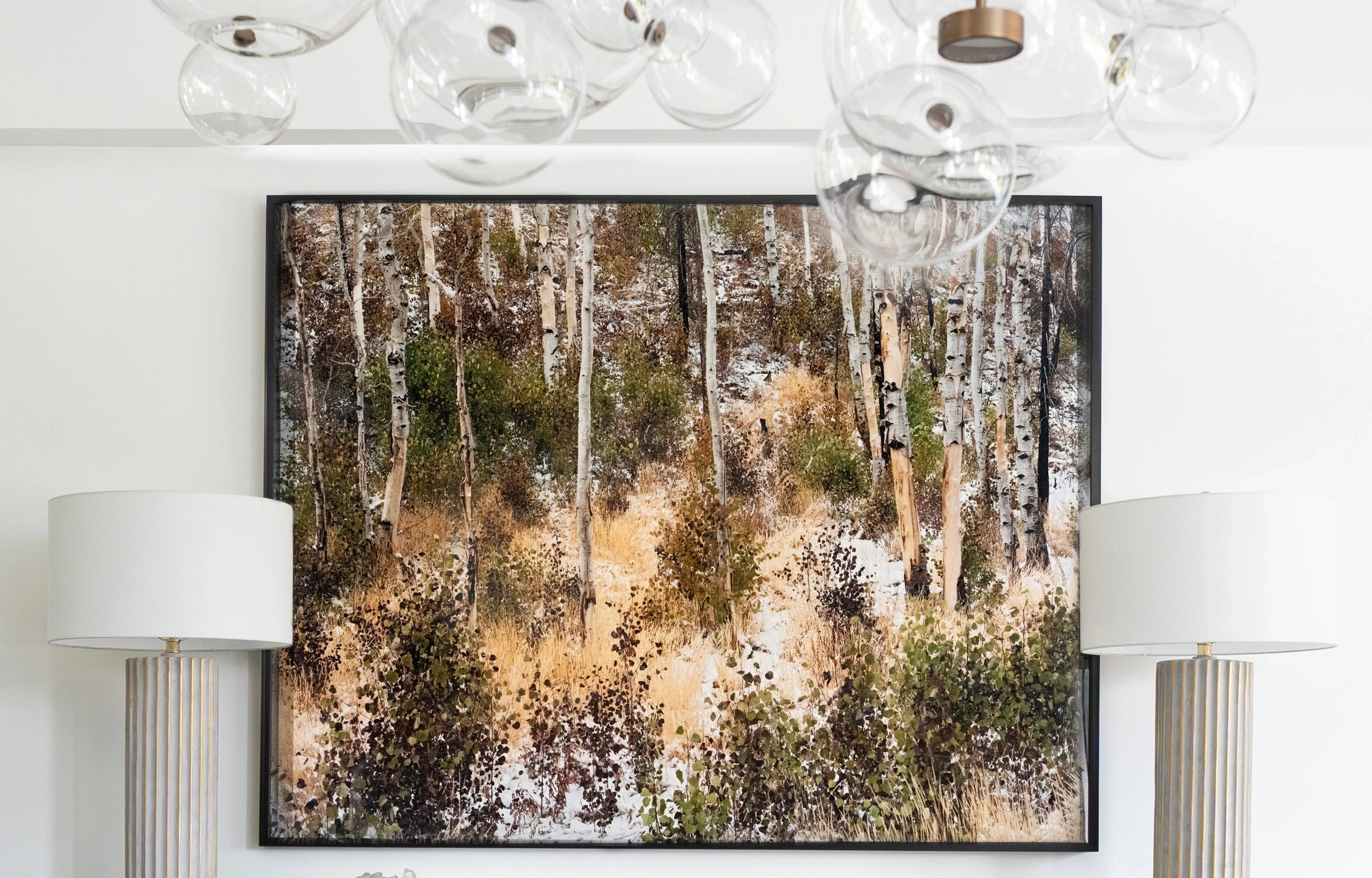 A photograph, flanked by two identical lamps, hangs above University House's dining table. The photograph depicts thin white aspen trunks surrounded by yellow grasses and green bushes.